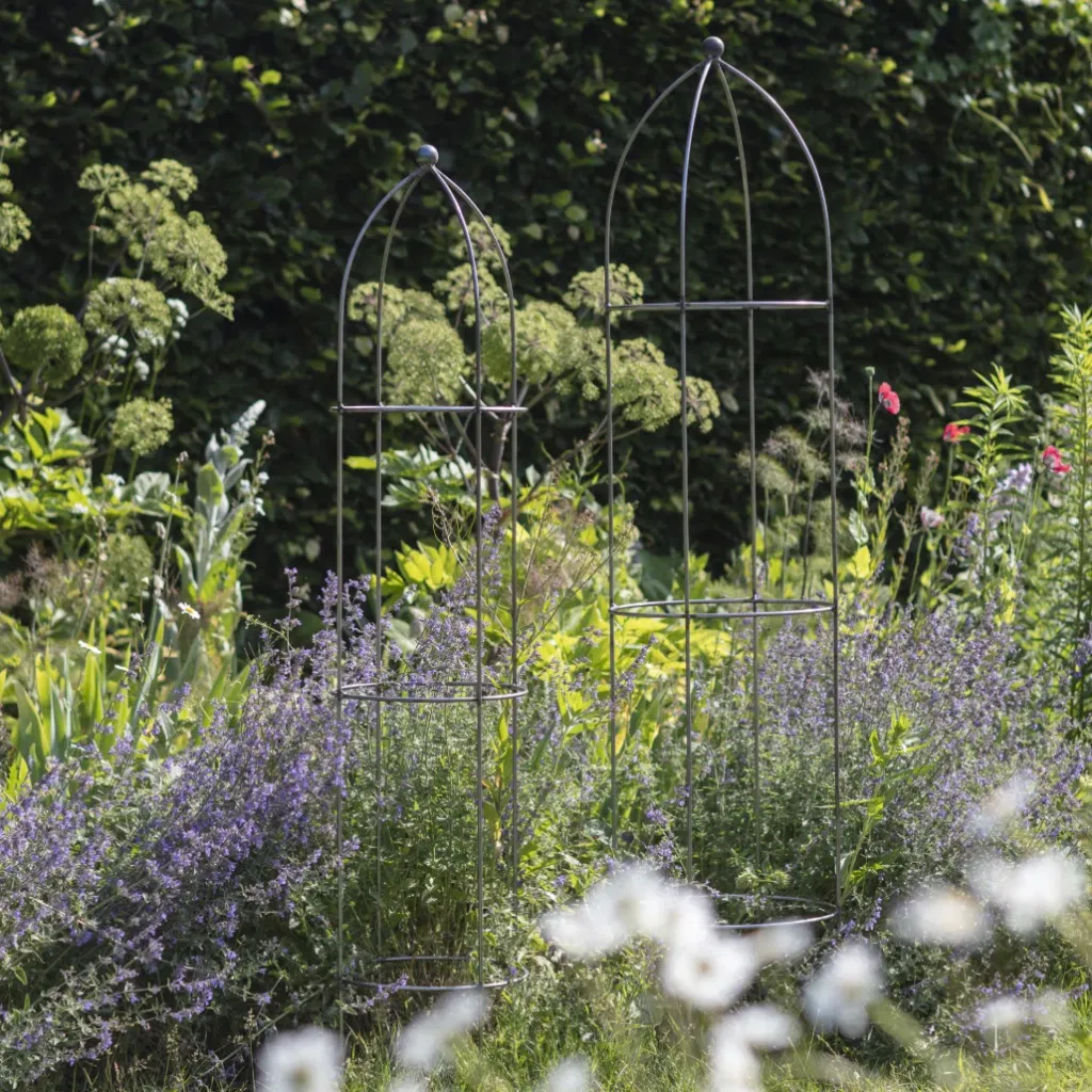 A pair of raw metal garden obelisks of different heights positioned among purple flowers and green shrubs.