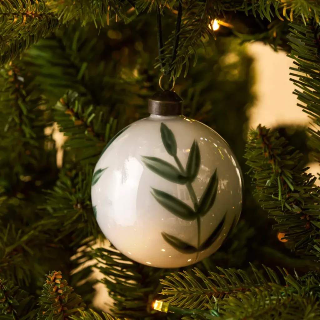 A single Bibury hand-painted glass rosemary Christmas bauble with a pearl finish hanging from a green pine branch.