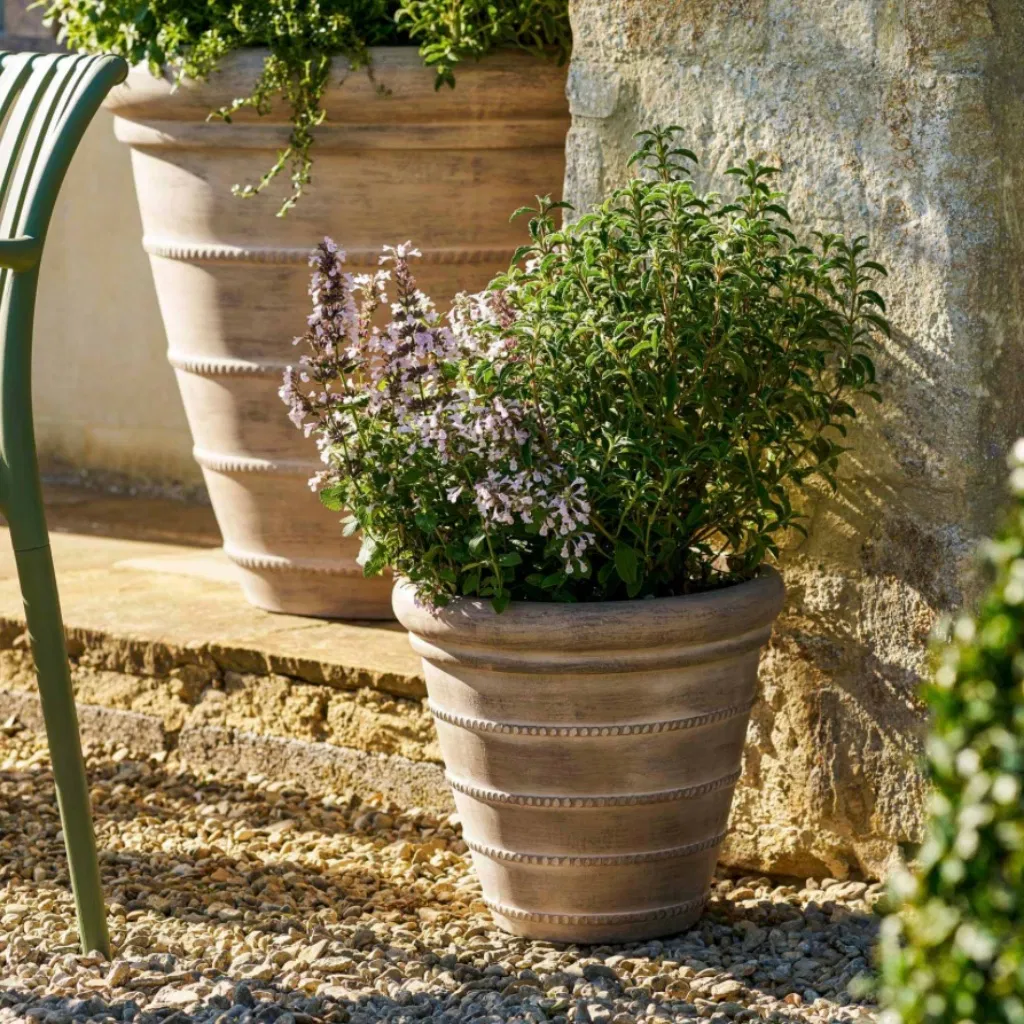 Close-up of the small 36cm Cotswold ribbed planter in washed terracotta ficonstone, planted with green herbs and pale purple flowers on a gravel patio.