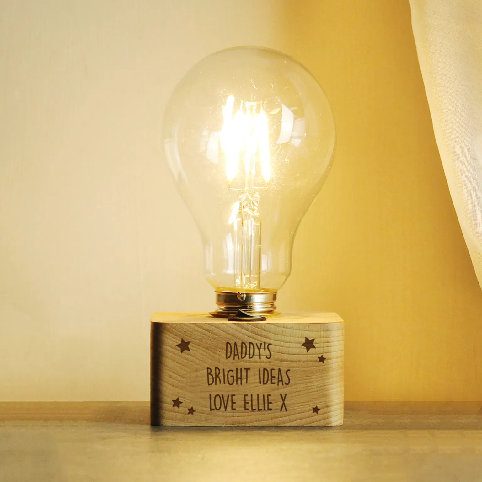 A personalised stars LED bulb table lamp by Abode Home & Gifts with a solid wooden base engraved with "DADDY'S BRIGHT IDEAS LOVE ELLIE X" lit up against a soft grey background.