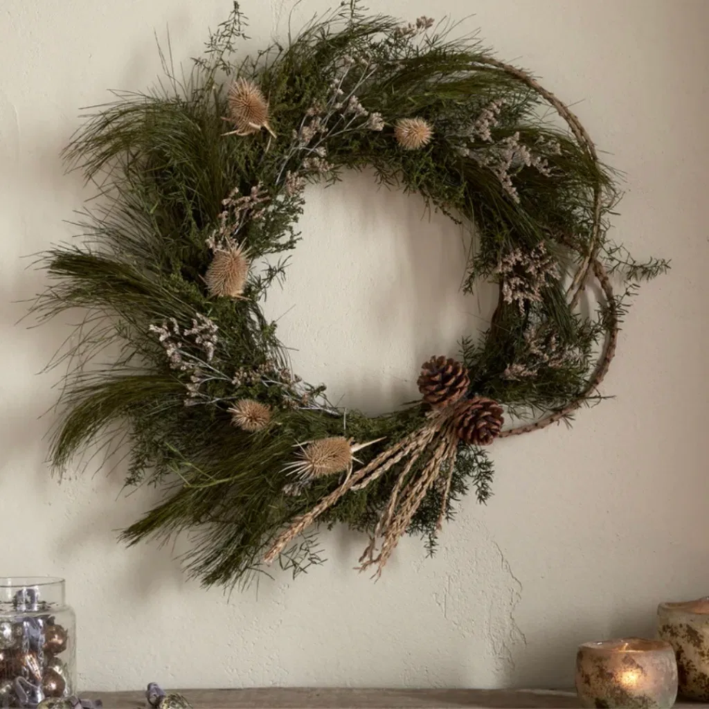 Elowen moss and thistle wreath hung on a neutral wall above a rustic wooden shelf.