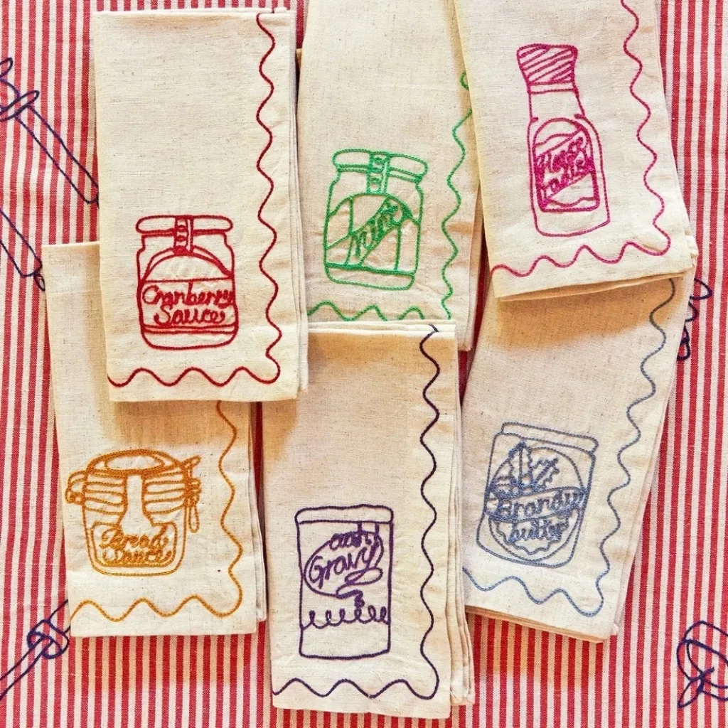 Set of six white cotton-linen napkins with colourful embroidered Christmas condiment jars and scalloped edges.