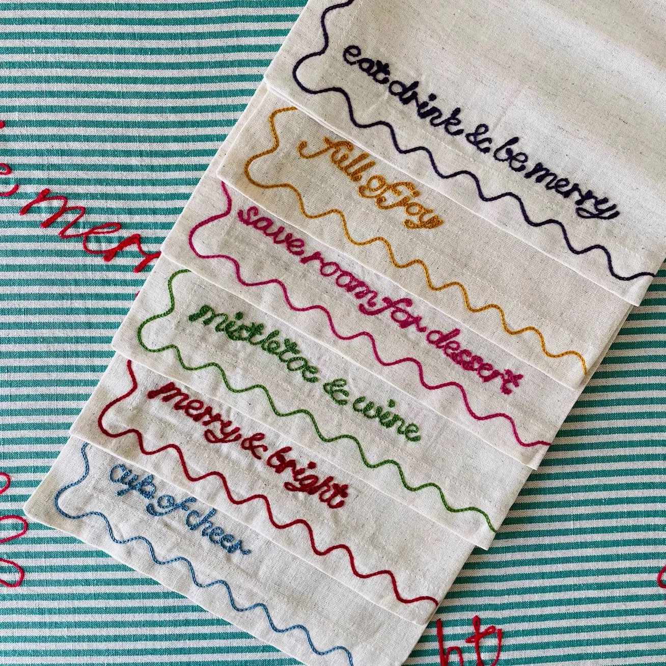 Set of six cotton-linen Christmas napkins with colorful embroidered phrases and scalloped edges.