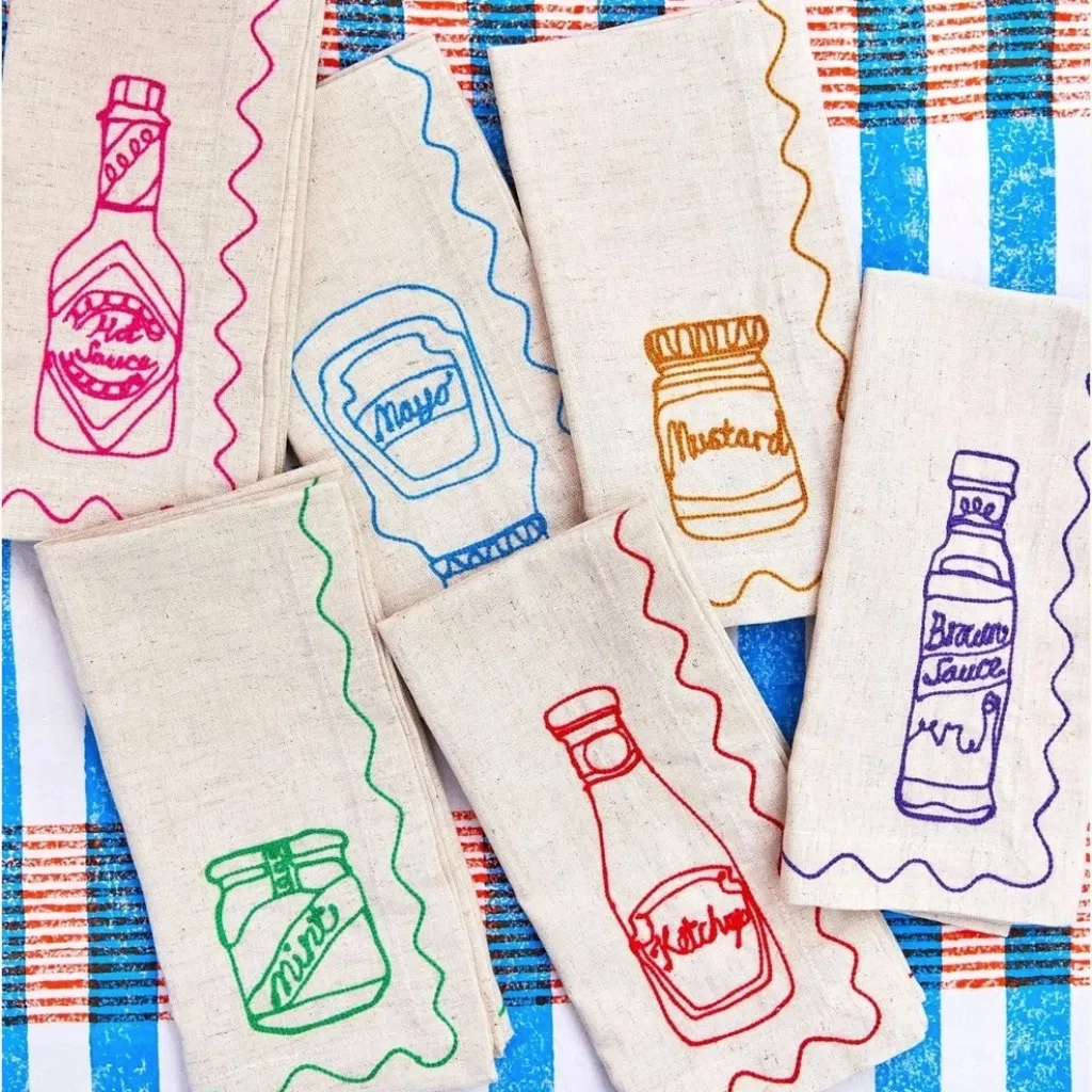 Six cream napkins with embroidered Ketchup, Mayo, Mustard, Hot Sauce, Brown Sauce, and Mint Sauce bottles on a blue striped background.