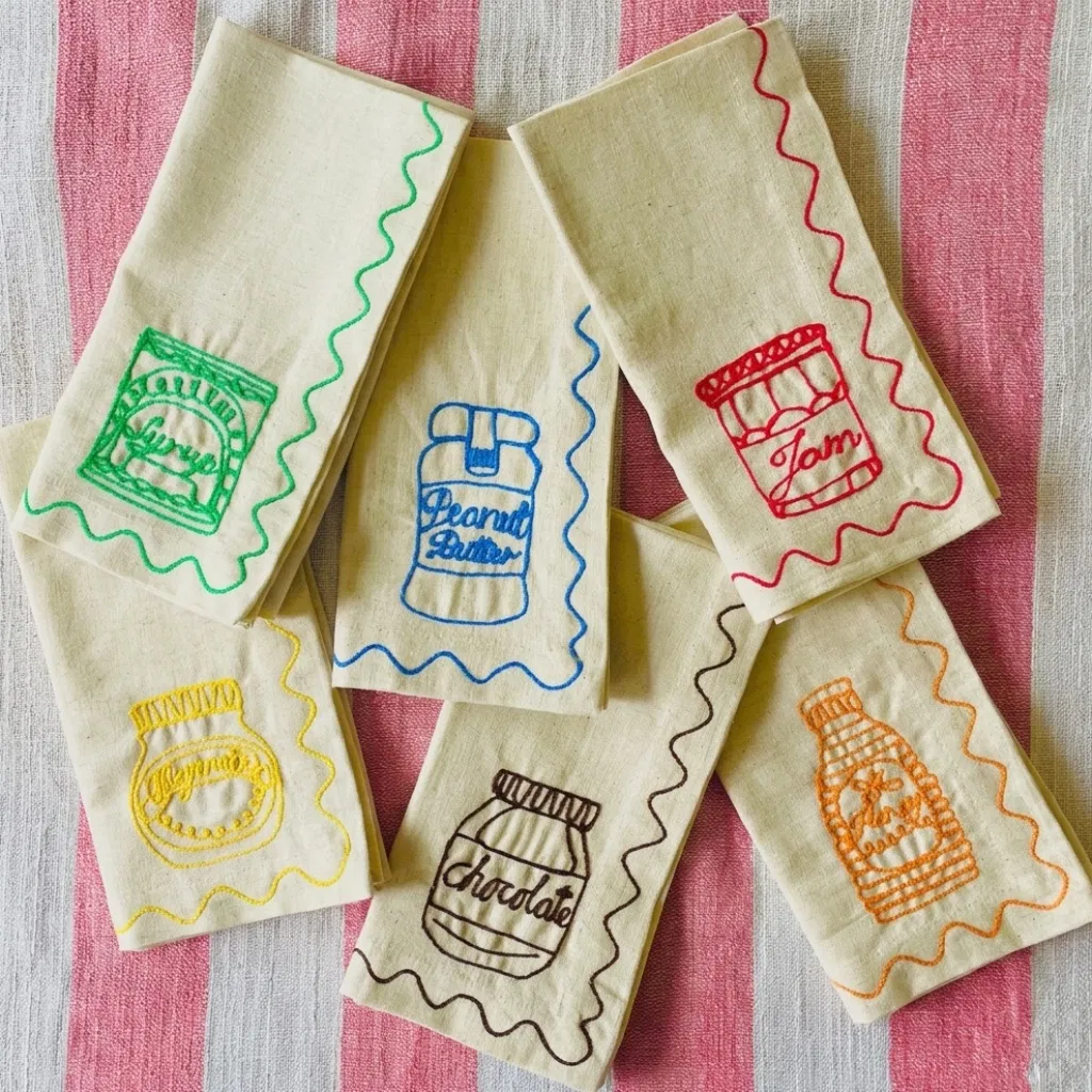 Six cream cotton-linen napkins with colorful embroidered jars of Jam, Peanut Butter, Honey, Marmite, Chocolate Spread, and Syrup on a pink striped background.