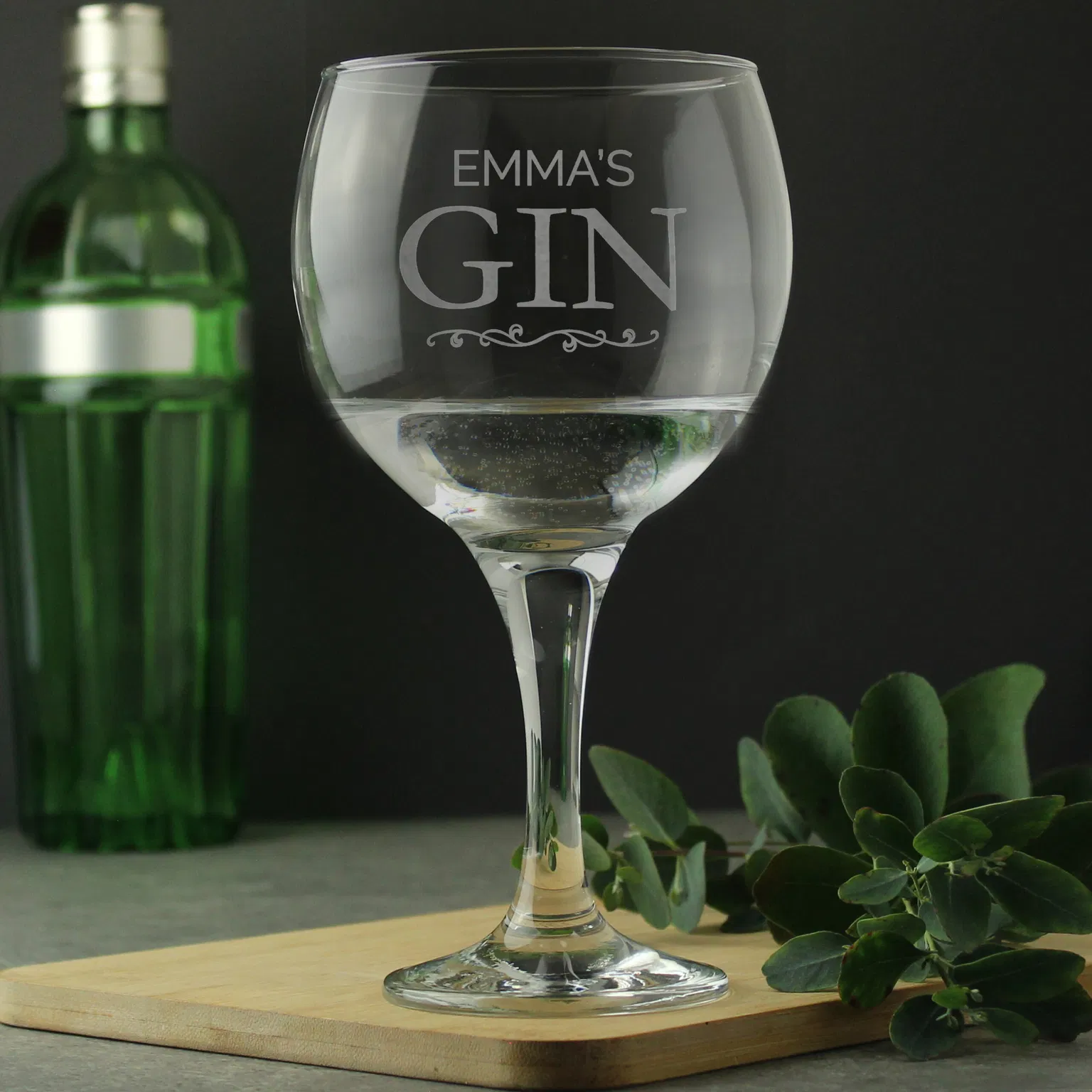 A personalised stemmed gin balloon glass by Abode Home & Gifts, featuring the custom laser-engraved text "EMMA'S GIN" above a decorative scroll, filled with a clear gin drink on a wooden serving board next to a green gin bottle and leafy eucalyptus sprigs.