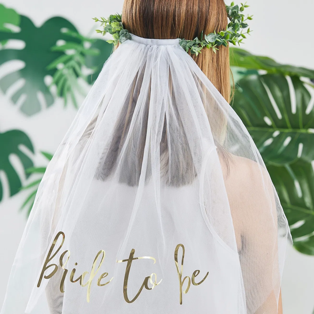 Rear view of a bride wearing a faux green eucalyptus foliage leaf crown headband with a flowing white tulle veil featuring gold-foiled cursive Bride to Be text.