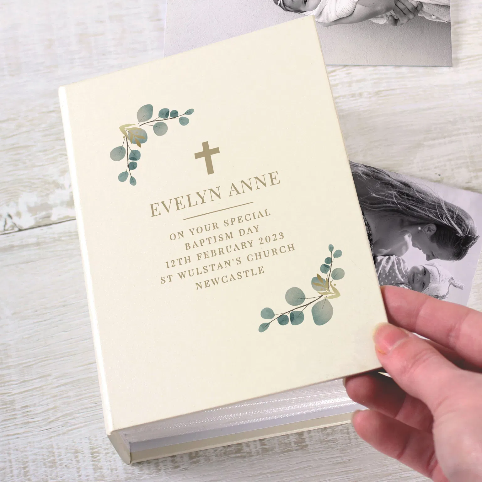 A hand holding an ivory hardback 6x4 photo album by Abode Home & Gifts next to black and white baby photos. The cover features a small cross, eucalyptus leaves, and text reading "EVELYN ANNE ON YOUR SPECIAL BAPTISM DAY 12TH FEBRUARY 2023 ST WULSTAN'S CHURCH NEWCASTLE".
