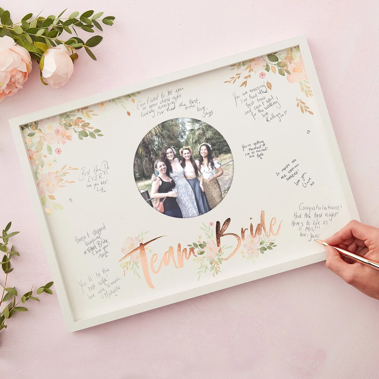 Top-down view of a white signature guest book frame with signed messages and a rose gold foiled Team Bride mount as a hand writes on it.