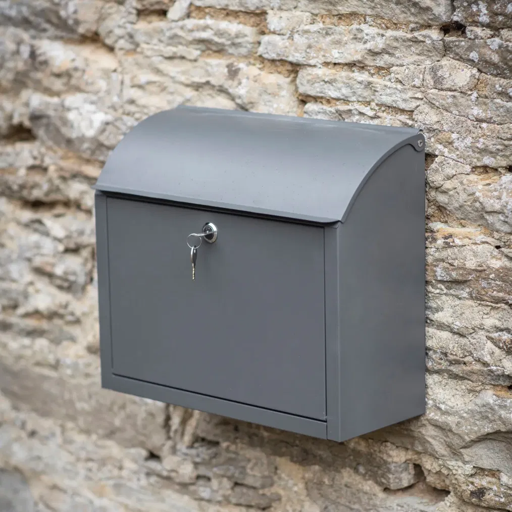 A charcoal grey metal parcel box mounted on a cream wooden wall with a secure lock and handle.