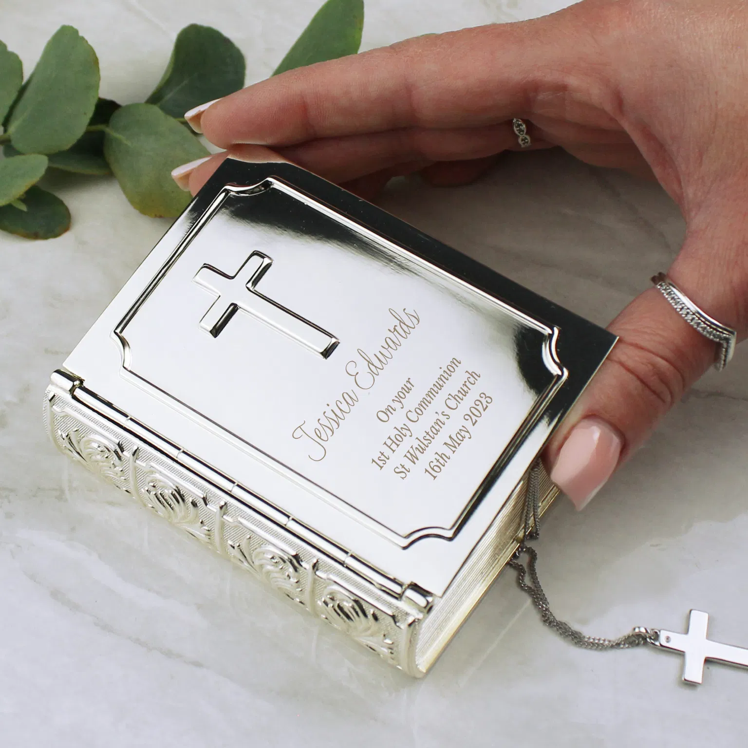A person's hand with silver rings holding a polished silver bible trinket box by Abode Home & Gifts on a white marble tabletop, engraved with "Jessica Edwards On your 1st Holy Communion St Wulstan's Church 16th May 2023", next to a silver crucifix.