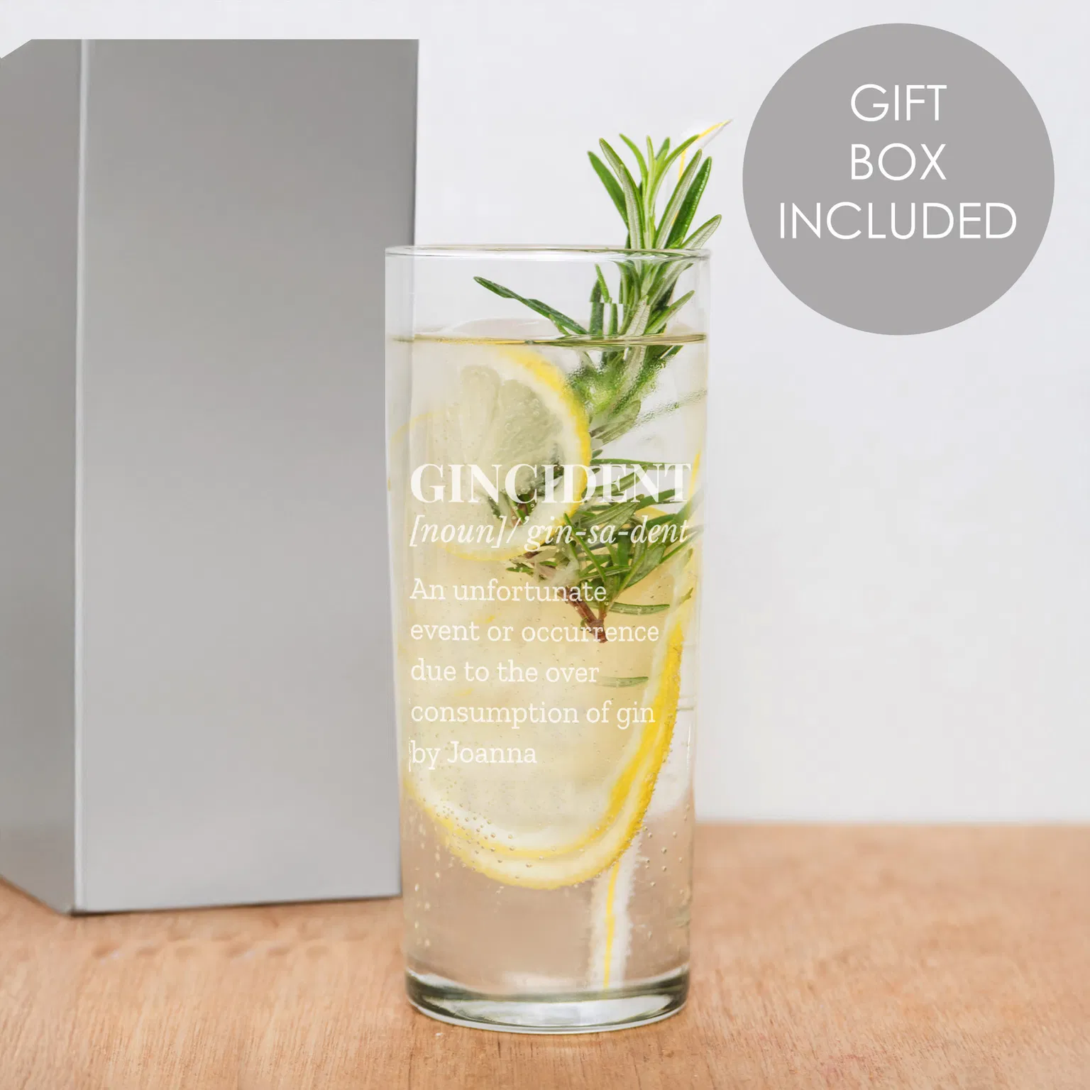 A tall, clear personalised highball glass by Abode Home & Gifts, filled with a gin drink, lemon slices, and a fresh rosemary sprig. The glass shows a frosted engraving of a "Gincident" dictionary definition ending with the name "Joanna", standing next to a grey gift box on a wooden board. A grey circular badge reads "Gift Box Included".