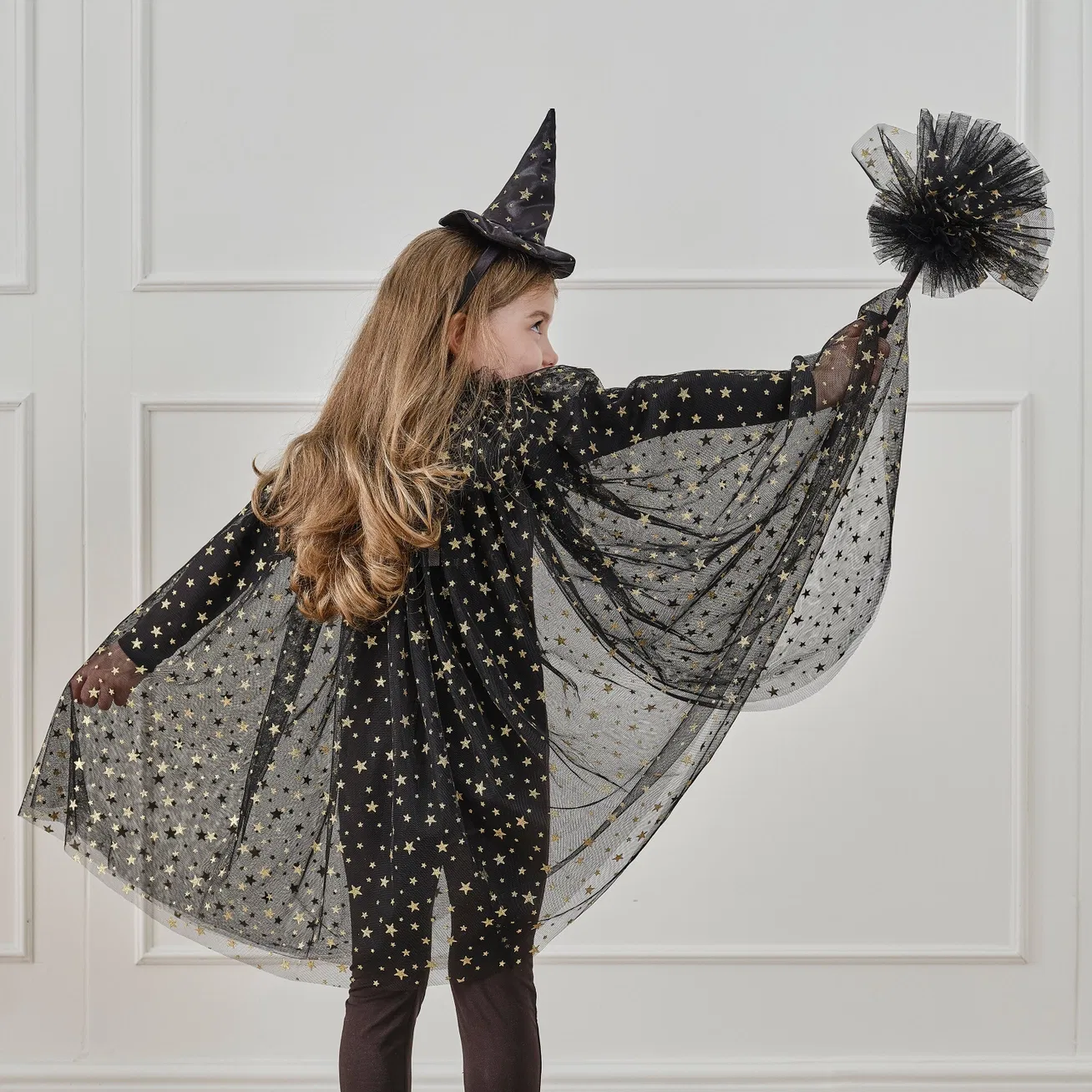 Back view of a child wearing a black tulle witch costume cape with gold foil stars and a matching witch hat headband.