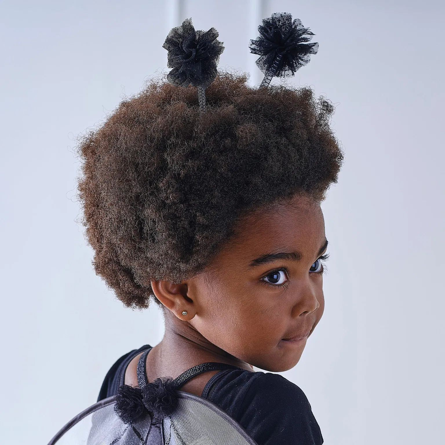 Over-the-shoulder portrait of a young girl with an afro wearing a bumble bee headband with black tulle pom pom antennae.