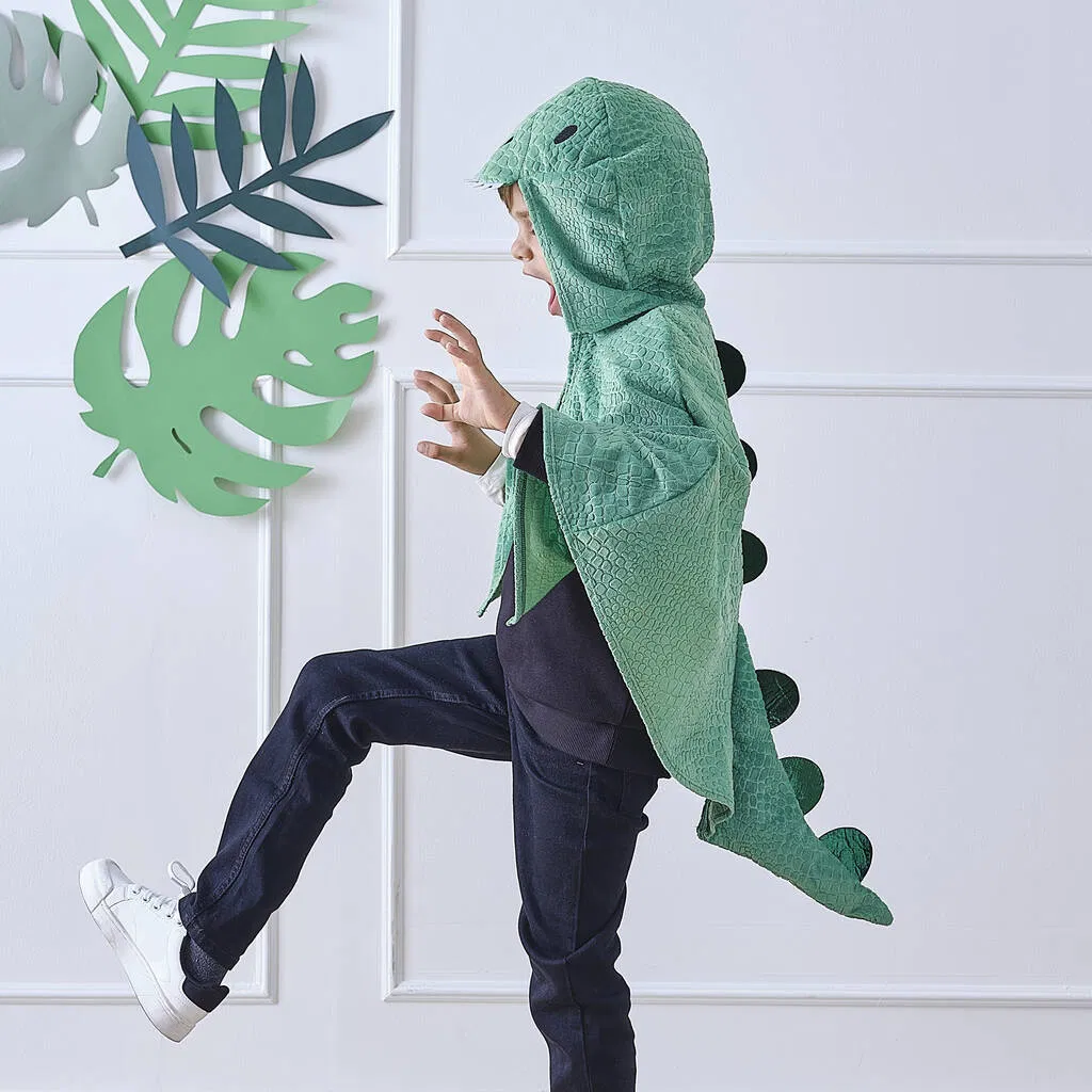 ide view of a child in mid-stride wearing a plush green dinosaur costume cape with scales, spines, and a character hood.