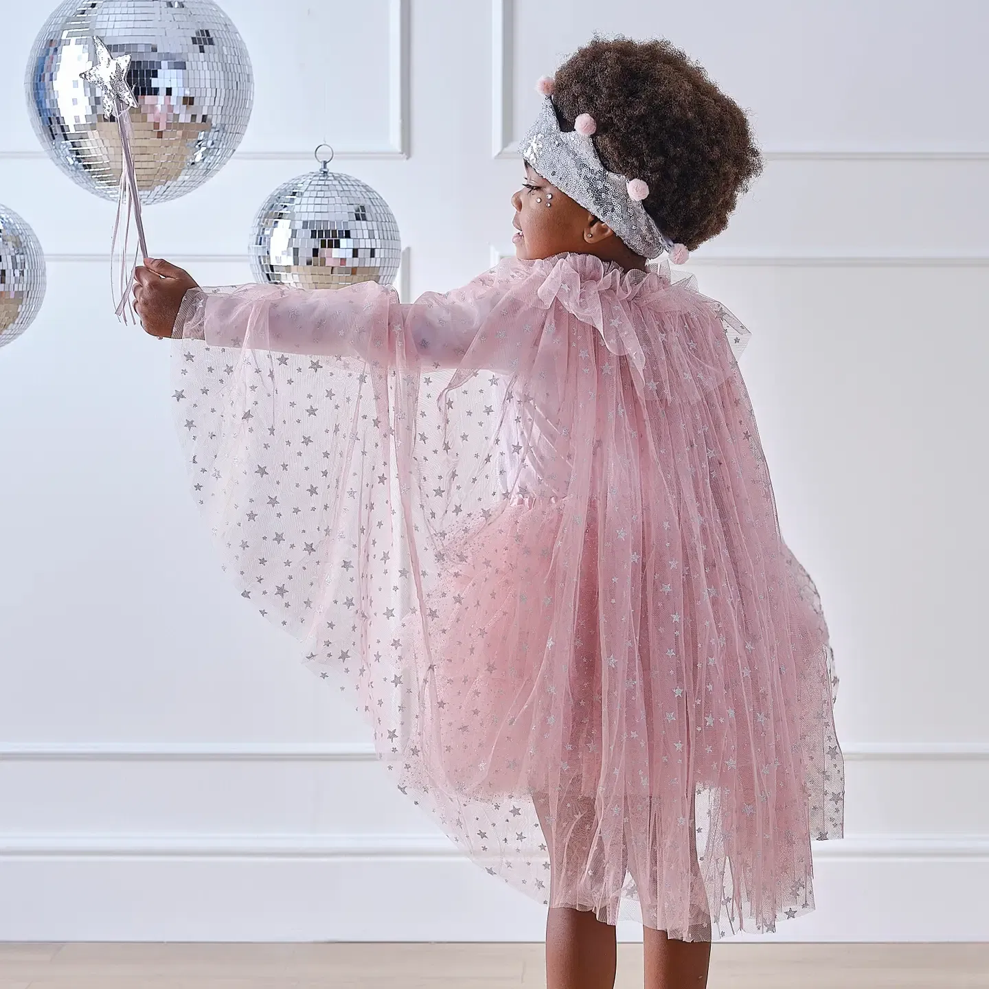 Back view of a child wearing a pink tulle fairy cape with silver stars while holding a silver wand next to disco balls.
