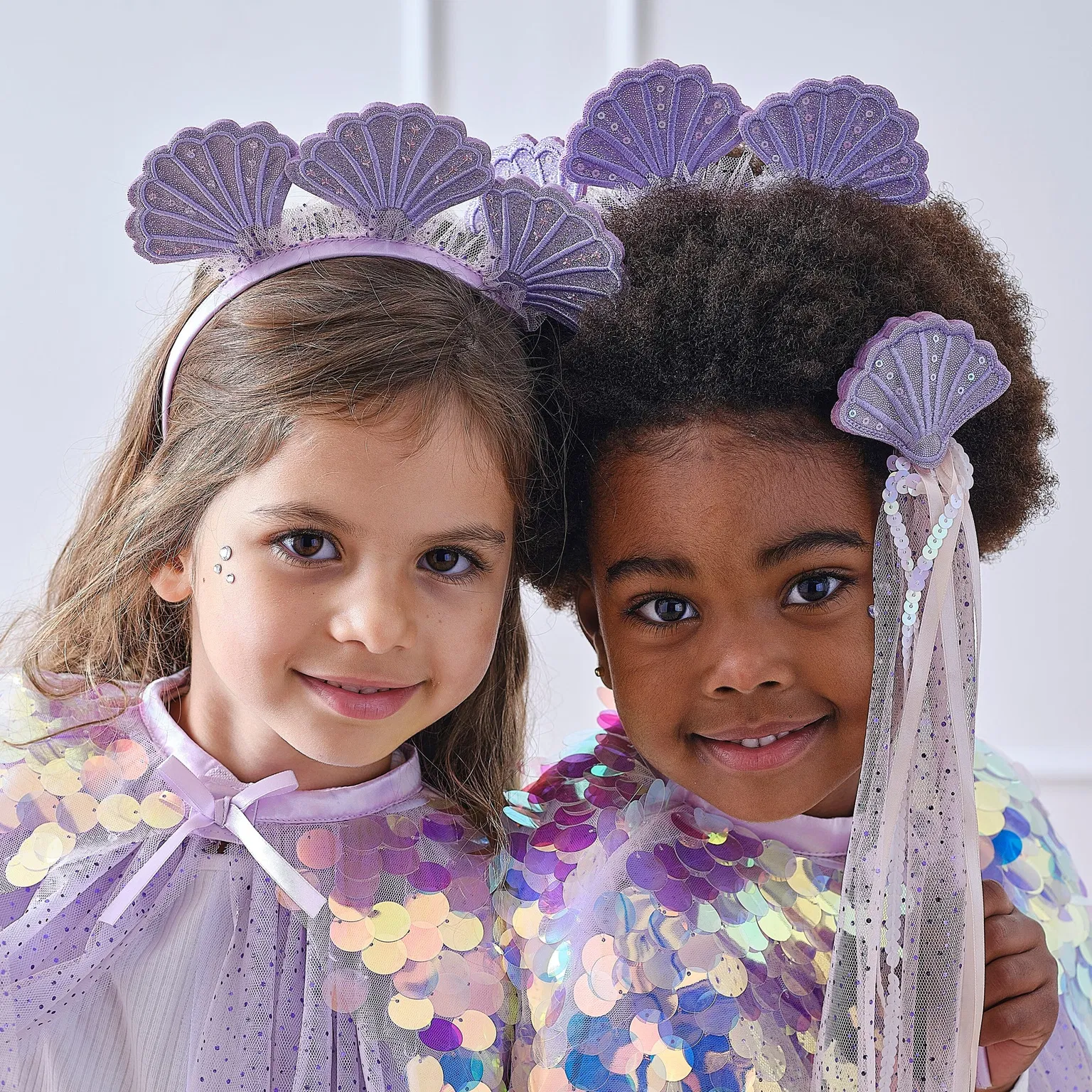 Close-up portraits of two young girls smiling while wearing matching purple shell mermaid headbands and tulle capes.