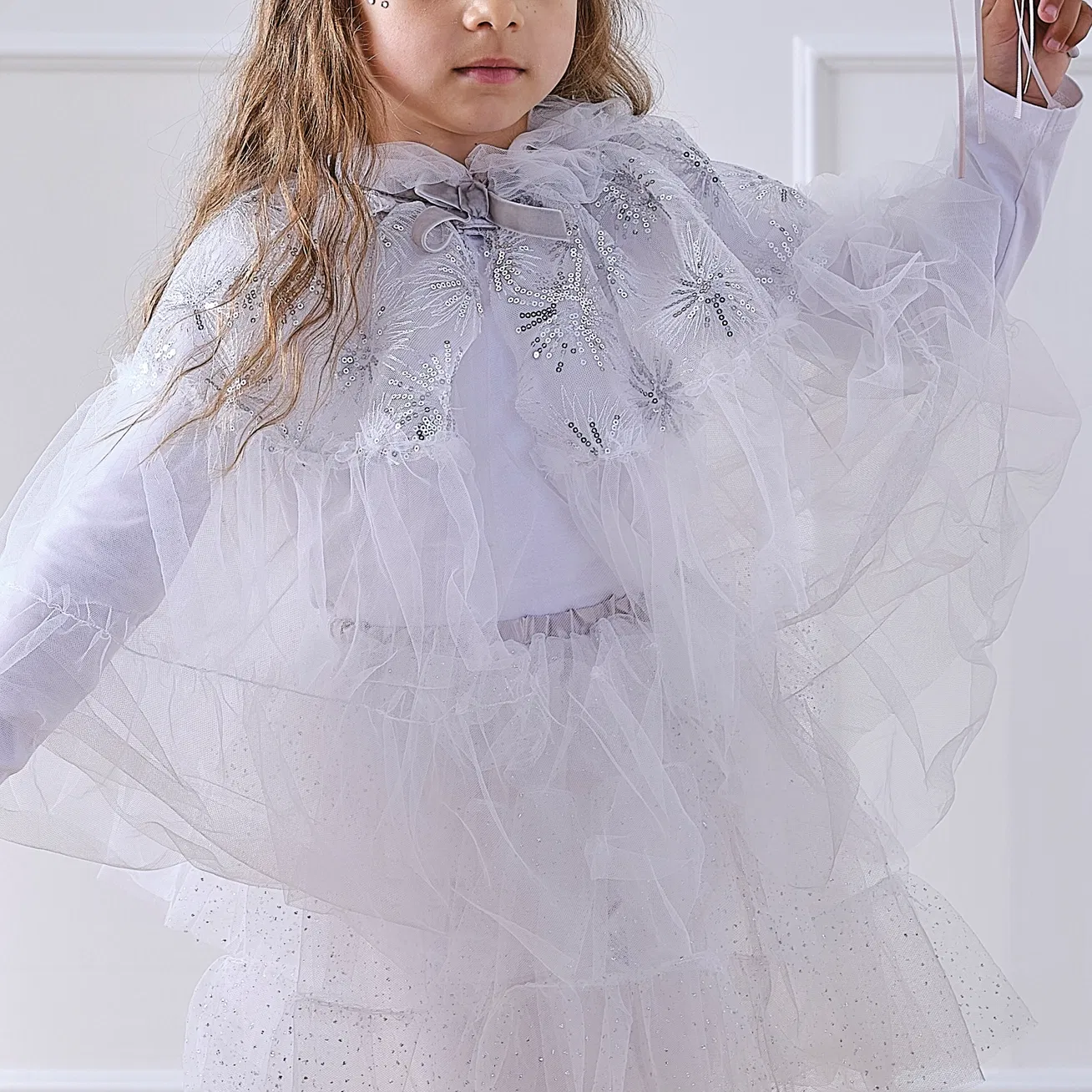 Close-up of a child wearing a white tulle winter fairy princess costume cape with silver sequin snowflake patterns and a silver velvet ribbon tie.
