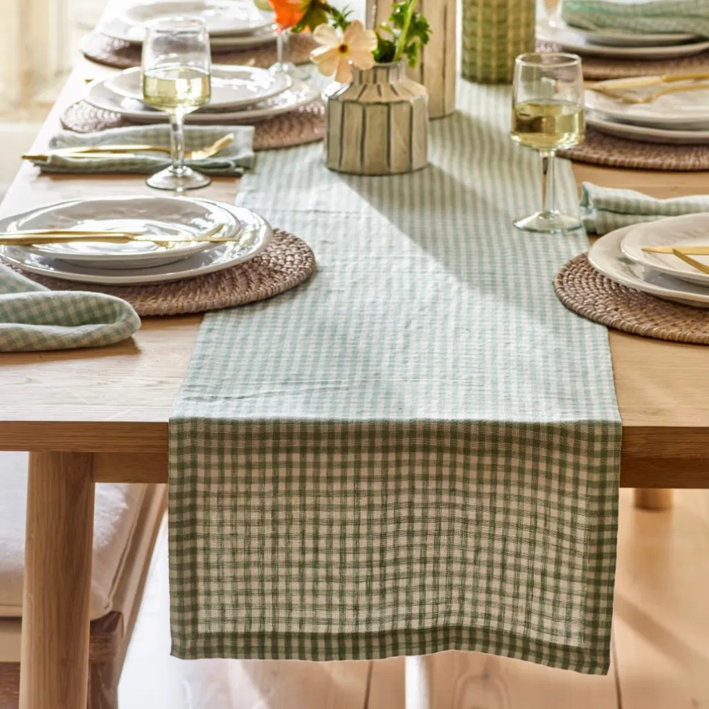 A rosemary green and cream checked linen table runner styled on an oak dining table with wine glasses and plates.