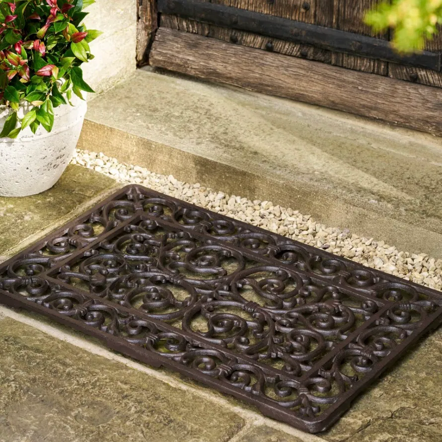 An ornate dark brown cast iron doormat placed on stone steps outside a rustic wooden door by Abode Home & Gifts.