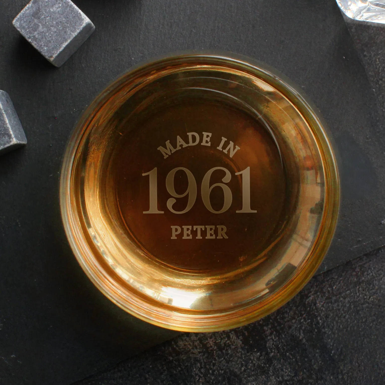 A top-down overhead shot looking straight inside a heavy glass tumbler by Abode Home & Gifts filled with golden whiskey. The bottom of the glass is custom base-engraved with the words "MADE IN 1961 PETER" in crisp capital letters. The glass sits on a square black slate coaster next to grey whiskey cooling stones.
