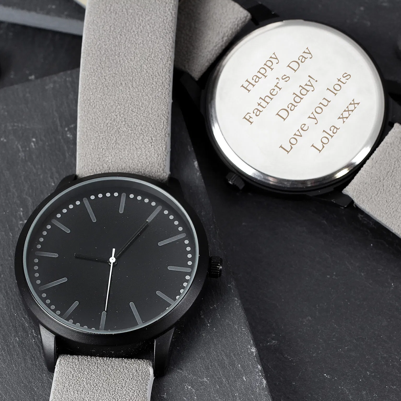 A personalised men's black watch by Abode Home & Gifts with a grey strap, showing the front face and the stainless steel back engraved with 'Happy Father's Day Daddy! Love you lots Lola xxx', styled on a slate surface.