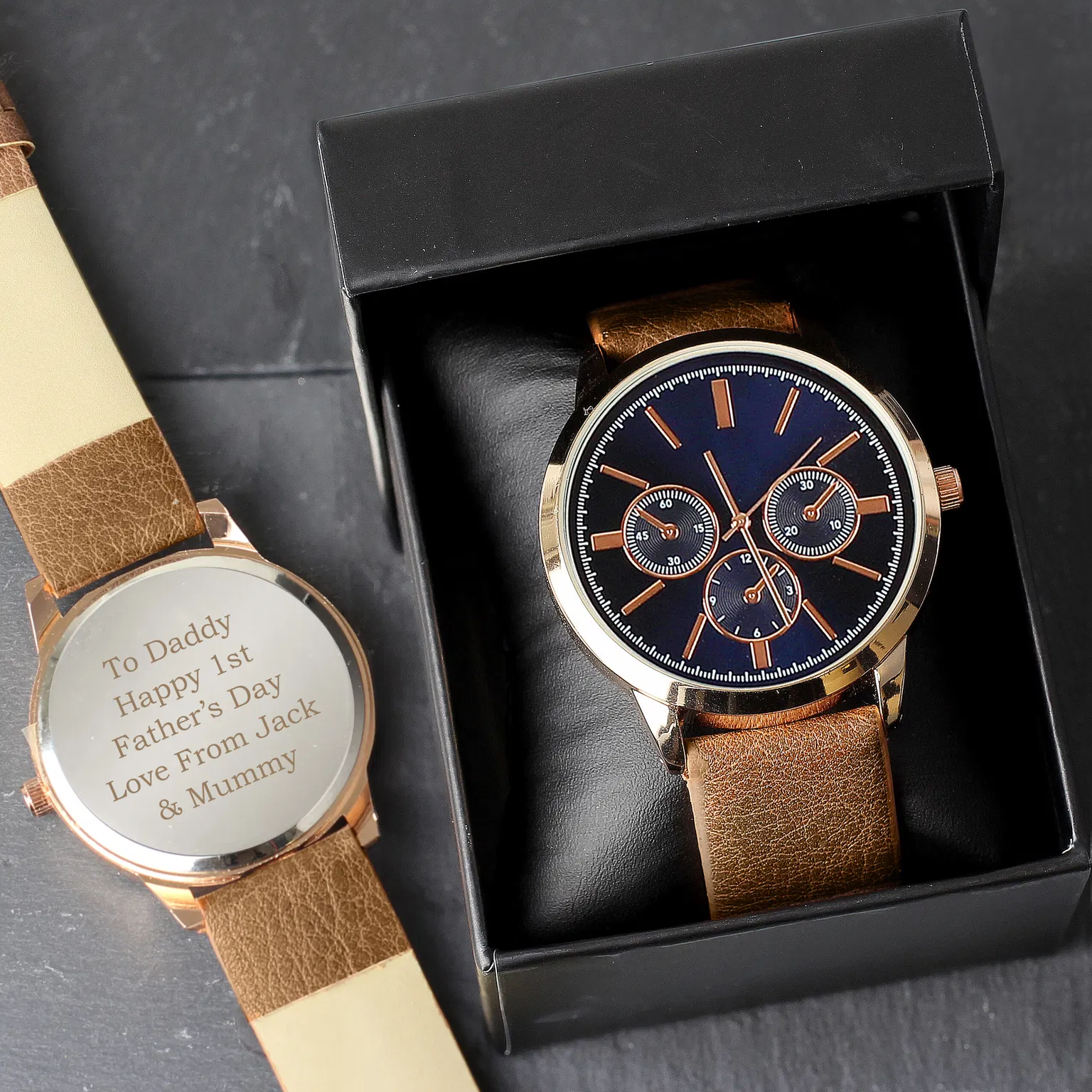 A personalised men's rose gold tone watch by Abode Home & Gifts with a blue decorative face and brown strap displayed in an open black gift box, next to a second watch showing the back engraved with "To Daddy Happy 1st Father's Day Love From Jack & Mummy" on a dark slate background.