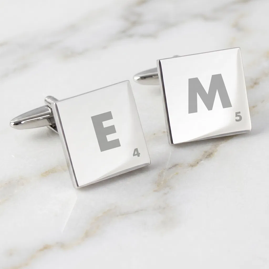 A pair of silver Scrabble tile cufflinks resting on a white marble surface, displaying the engravings 'E 4' and 'M 5'.