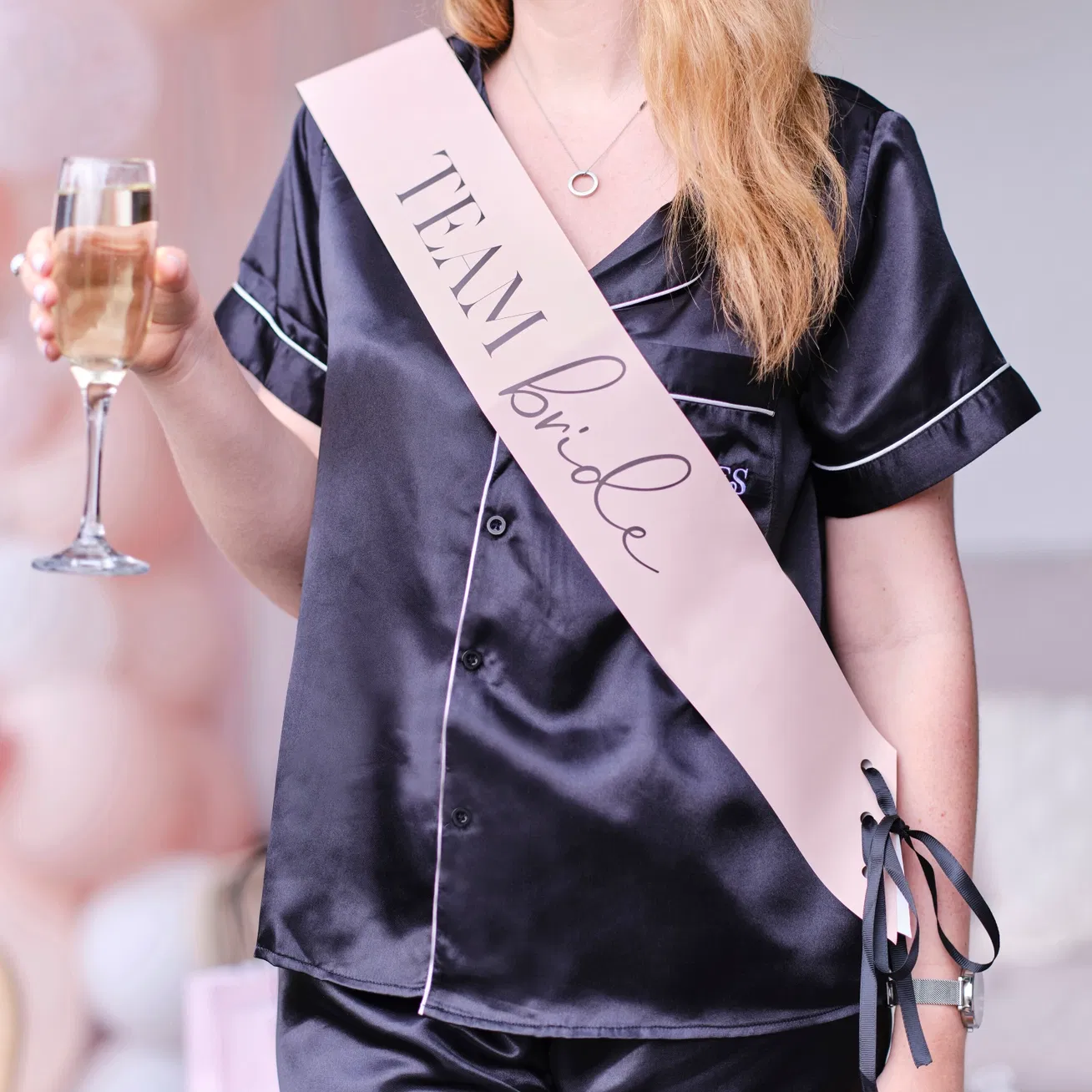Close mid-section view of a bridesmaid wearing a pale pink cursive script sash over a black satin pyjama set while holding a champagne glass.