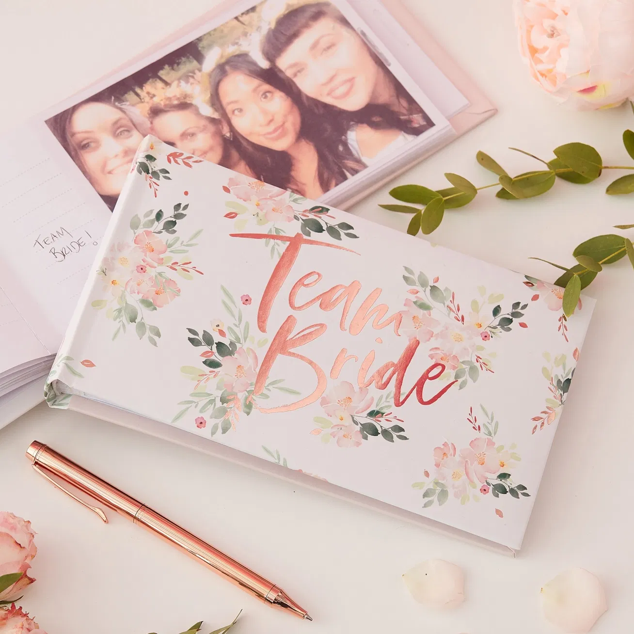 Rose gold foiled Team Bride floral hen party photo album lying flat beside a rose gold pen and green foliage.