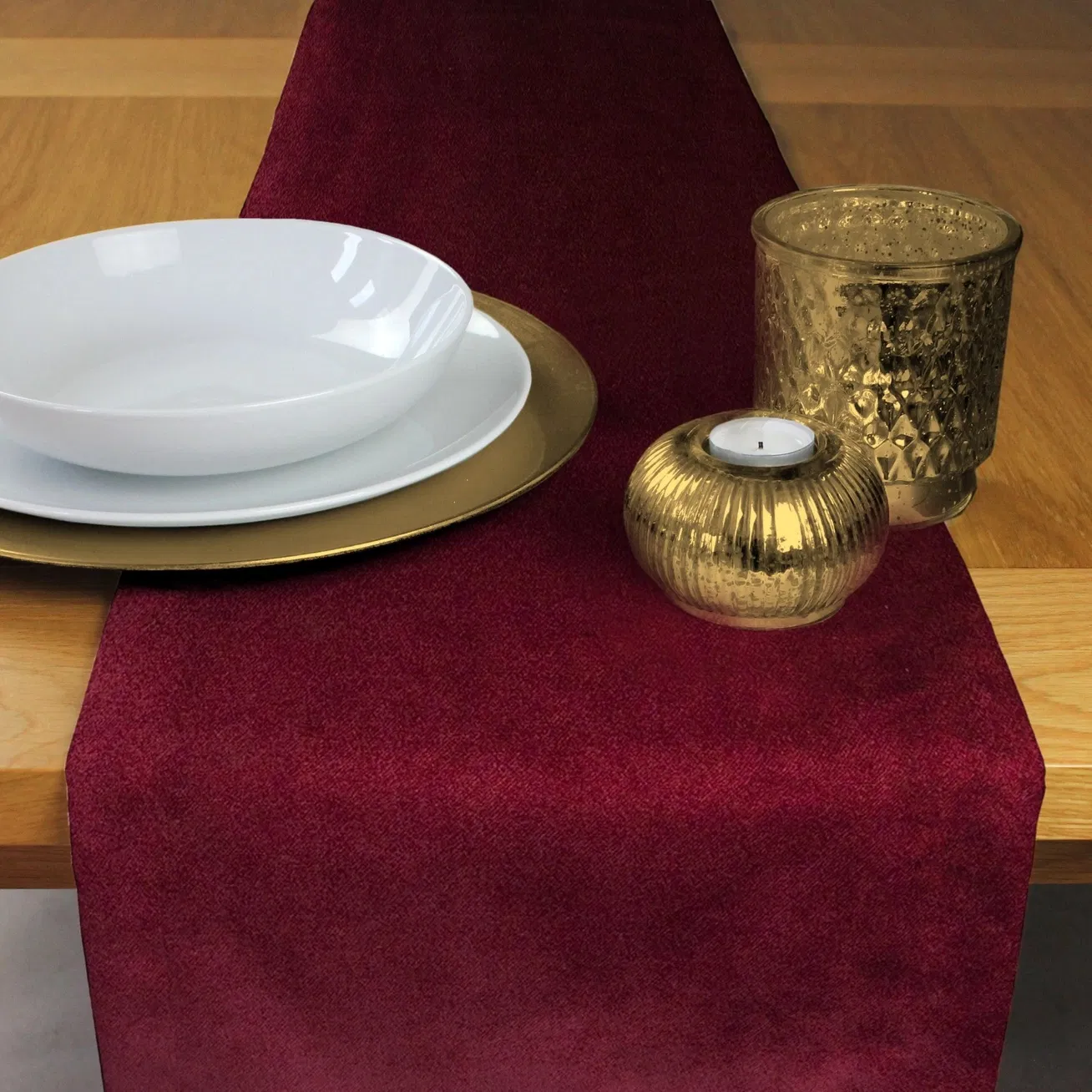 Luxury wine red matt velvet table runner styled with gold candle holders and white dinnerware on a wooden table.